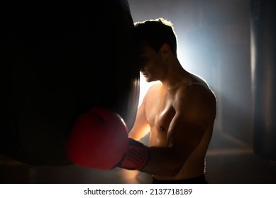 Tired boxer leaning on punching bag. Sweaty shirtless man in boxing gloves standing in gym against light. Sport and endurance concept - Powered by Shutterstock