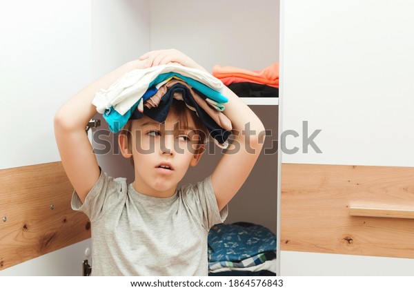 Tired boy holding\
stack of clothes on his head. Kid organizing clothes in wardrobe.\
Order in the closet. Wardrobe with child\'s clothing. The boy puts\
things in order in the\
closet.