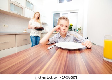 Tired Boy Sitting At The Table Waiting For Dinner And Playing With Forks While His Mother Cooking At The Kitchen.