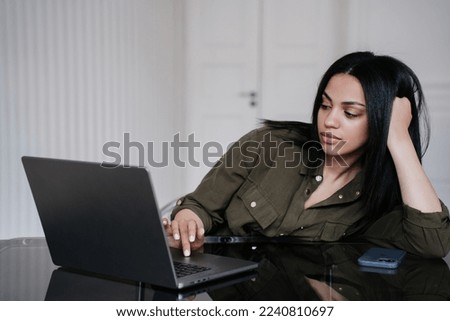 Tired brazilian young woman sitting at table with laptop at office, typing, exhausted by working all day long.  African american student leaning on hand, sleepy. Overloaded businesswoman relaxing