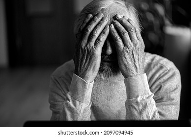 A tired, broken, resigned, preoccupied elderly man sits in front of a laptop, computer and covers his face with his hand. 