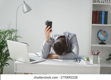 Tired Business Man In Gray Shirt Sitting At Desk Work On Laptop Pc Computer In Light Office On White Wall Background. Achievement Business Career Concept. Put Head On Table Hold Cup Of Coffee Or Tea