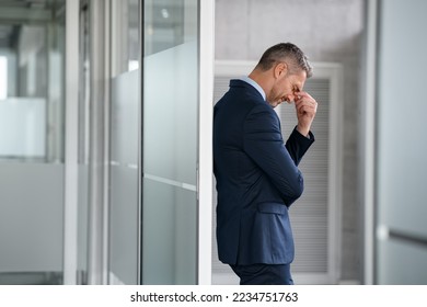 Tired business man suffering from headache at work. Mid man entrepreneur feeling stressed while pressing eye standing and leaning at glass wall in office. Depressed businessman suffering from migraine - Powered by Shutterstock