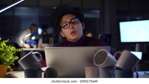 Tired Businessman Falling Asleep At Table With Computer And Coffee Cup During Night Time In Office. Exhausted Asian Male Employee With Messy Hair Sleeping At Desk In Dark Office