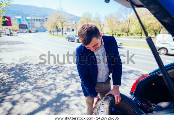 A tired\
businessman on a sunny day is taking a tire out of his car in order\
to be able to change his flat\
tire.