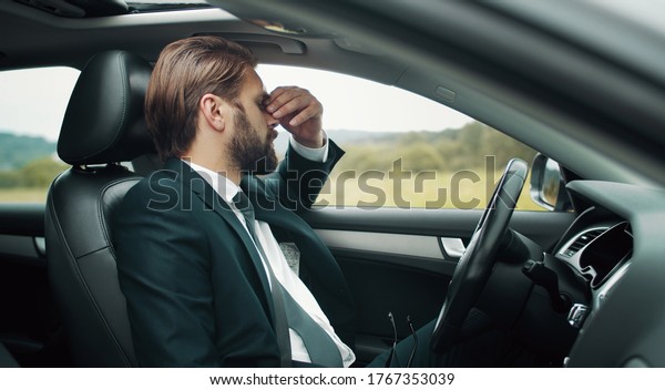 Tired businessman\
sitting on driver\'s seat touching bridge of nose staying somewhere\
in countryside