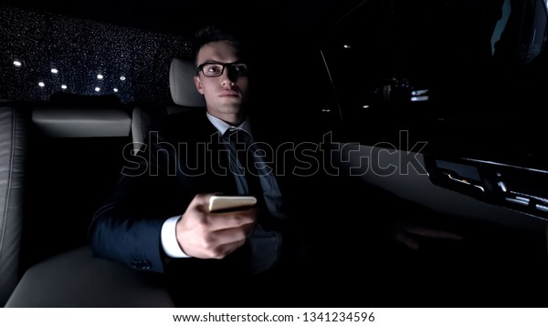 Tired businessman surfing net on smartphone,\
riding in car, analyzing\
project