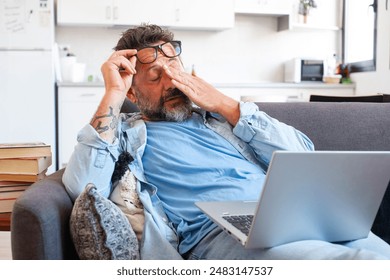 Tired businessman takes off glasses and feels eye strain from working online with laptop, Caucasian man sits on sofa and rubs his eyes, feels headache and lack of energy - Powered by Shutterstock