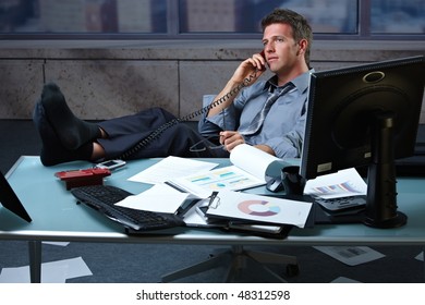 Tired businessman taking break speaking on landline phone with shoes off feet up on office desk holding glasses.