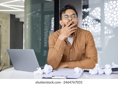 Tired businessman yawning at his desk surrounded by a laptop and crumpled papers in a modern office setting. Concept of fatigue, overworked, and stress. - Powered by Shutterstock