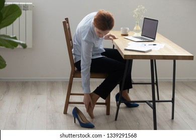 Tired businesswoman sitting on office chair and massaging foot, feeling discomfort after wearing shoes with heels, tense muscle, employee doing easy exercise at workplace