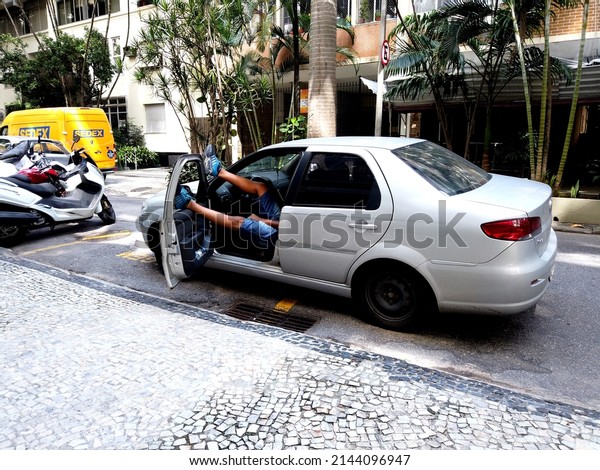 Tired car driver, sleeping in the car with his\
feet over the door. Lazy man wearing blue shoes, sleeping on the\
road waiting, shaking his foot. Gray Fiat Siena car. Rio de\
Janeiro, Brazil. 04.08.22