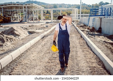 Tired Caucasian Handsome Worker In Overall Walking Outdoors, Holding Helmet And Wiping Sweat. In Background Is Oil Tank.