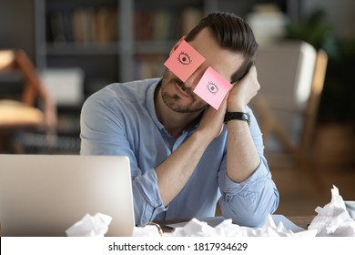 Tired Caucasian male worker fall asleep at workplace in office, have sticker pads on eyes. Exhausted young man take nap doze off sleep at desk, overwhelmed with work. Fatigue, exhaustion concept.