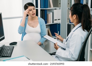 Tired Chinese Expectant Patient Is Touching Her Forehead While Telling Her Doctor With A Clipboard About Her Dizziness During Pregnancy Checkup In The Clinic.