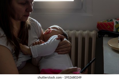 Tired Concerned Mother Rocking Sleeping Baby In Kitchen During The Day At Home. New Mother Looking At Smatphone While Rocking Her Baby. Portrait Of Young Woman And Cute Little Child In Home Interior.