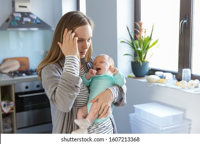 Tired Concerned New Mother Rocking Crying Baby In Kitchen. Portrait Of Young Woman And Cute Little Child In Home Interior. Motherhood Concept