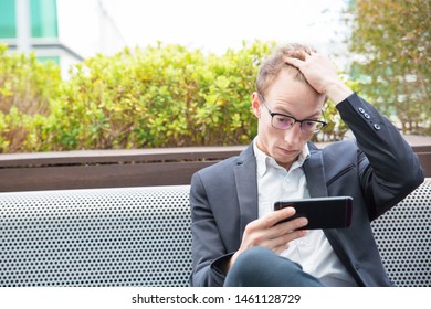 Tired Concerned Office Employee Using App On Mobile Phone. Young Business Man In Eyeglasses And Jacket Sitting Outside, Looking At Cellphone Screen And Scratching Head. Smartphone Using Concept