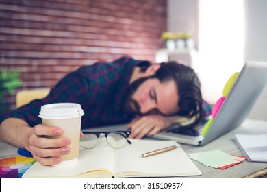 Tired creative editor holding disposable cup while sleeping on office desk