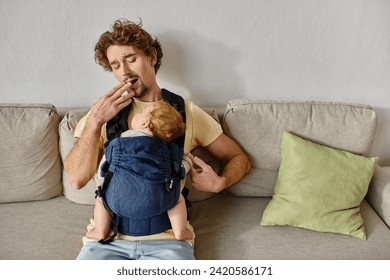 tired curly haired man yawning with infant son in baby carrier in living room, fatherhood and love - Powered by Shutterstock