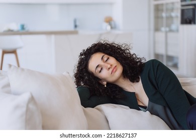 Tired Curly-haired Young Woman Fell Asleep Sitting On The Couch At Home. Portrait Of A Sleeping Young Spanish Woman Against The Background Of A Blurred Kitchen. Your Girlfriend Relax After A Day Home.