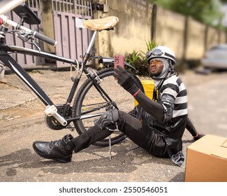 A tired delivery bike man sits on the ground,multitasking as he tries to fix his bike while checking his phone. The scene captures the resilience of a courier amid the challenges of urban Lagos street - Powered by Shutterstock