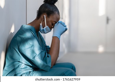 Tired depressed female african scrub nurse wears face mask blue uniform gloves sits on hospital floor. Exhausted sad black doctor feels burnout stress of corona virus frontline protection pray at work - Shutterstock ID 1761627959