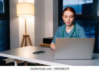 Tired Depressed Redhead Young Female Doctor In Blue Green Medical Uniform Working Typing On Laptop Computer Sitting At Desk In Dark Hospital Office Room Near Window At Night.