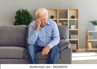 Tired, depressed senior man sitting on couch in living room feeling hurt and lonely. Aged, white-haired man touching forehead suffering from severe headache or recalling bad memories - Powered by Shutterstock