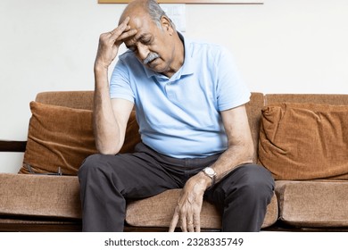 Tired, depressed senior man sitting on couch in living room feeling hurt and lonely. Aged, white-haired man touching forehead suffering from severe headache or recalling bad memories - Powered by Shutterstock