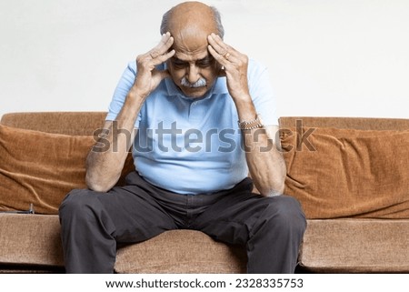 Tired, depressed senior man sitting on couch in living room feeling hurt and lonely. Aged, white-haired man touching forehead suffering from severe headache or recalling bad memories