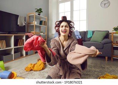 Tired Desperate Busy Housewife Cleaning Up Messy House. Unhappy Young Woman In Hair Curlers And Skincare Face Mask Picking Piles Of Clothes And Complaining About Mess And Chaos. Women's Chores Concept