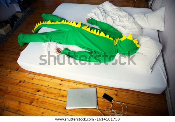 Tired dinosaur man lying face down in a simple bed,\
enjoying the creature comforts while gadgets charge on the floor\
beside him