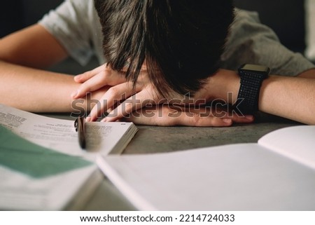 Tired disappointed teenage boy lowering his head sitting among pile of books, textbooks, school exercise books. Online learning, reading difficulties, education concept