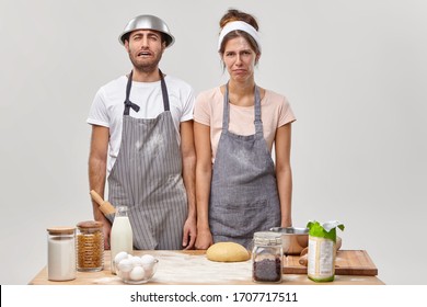 Tired Discontent Housewife And Husband Stand Together At Kitchen Near Table With Ingredients, Make Dinner For Family, Prepare Dough For Baking Pizza, Exhausted Of Routine Activity, Wear Aprons