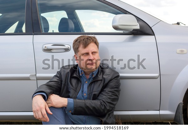 A tired driver sits on the grass near the car in\
the steppe.