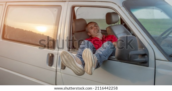 Tired driver sleeping inside of vehicle cabin\
stretching legs out open side window. Resting and breaking during\
hard road trip.