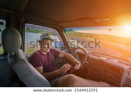 Tired driver wearing hat resting after work inside of truck cabin with digital tablet in hands