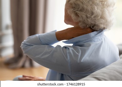 Tired Elderly 70s Woman Sit On Sofa Massaging Stiff Sore Neck Tensed Muscles Reduces Painful Feelings, Shoulder Back Pain Fibromyalgia Osteoarthritis Senile Chronic Diseases Concept Close Up Rear View