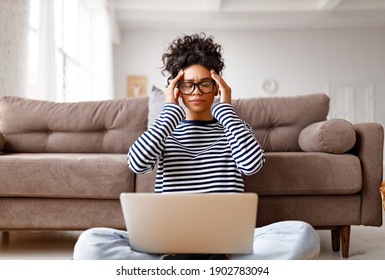tired ethnic woman with glasses touching head and closing eyes resting while sitting cross legged near laptop and working on remote project in cozy living room at home