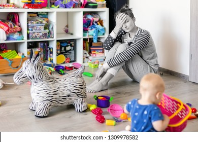 Tired Of Everyday Household Mother Sitting On Floor With Hands On Face. Kid Playing In Messy Room. Scaterred Toys And Disorder. Happy Parenting.