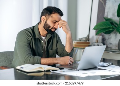 Tired exhausted arabic or indian man, office worker, manager or freelancer, sitting at his desk, tired of working in a laptop, overworked, having a headache, closed his eyes, needs rest and break