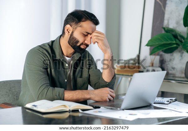 Tired exhausted arabic or indian man, office\
worker, manager or freelancer, sitting at his desk, tired of\
working in a laptop, overworked, having a headache, closed his\
eyes, needs rest and\
break