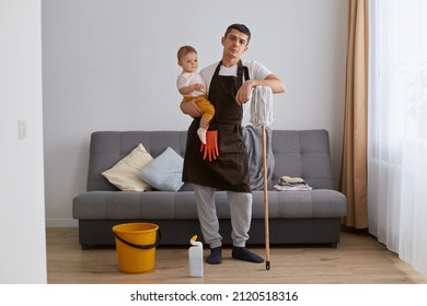 Tired Exhausted Brunette Young Adult Man Wearing Casual Attire And Brown Apron Cleaning House With Baby In Hands, Looking At Camera With Sad Upset Facial Expression.