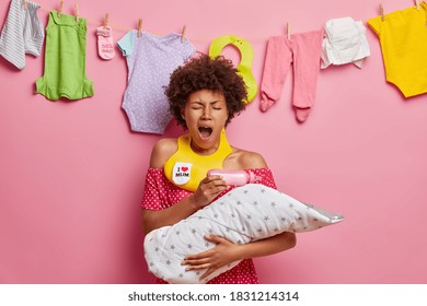 Tired exhausted ethnic mother yawns and wants to sleep feeds baby infant from milk bottle late at night takes care of newborn poses indoor against rope with washed clothing. Motherhood concept