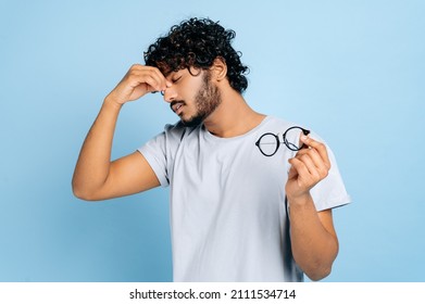 Tired exhausted indian or arab guy in a t-shirt, took off his glasses, closed his eyes, massages the bridge of his nose, has a migraine, headache, stands on an isolated blue background