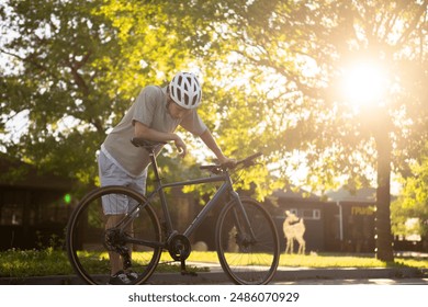 tired and exhausted man in a helmet takes a break from cycling in the park. He wears a grey shirt and white shorts, leaning on his black bike, surrounded by green grass and trees - Powered by Shutterstock