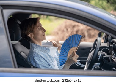 Tired exhausted middle aged woman waving blue fan suffers from stuffiness driving car on summer hot weather. Overheating, sultriness, high temperature, swelter in car with broken air conditioner.