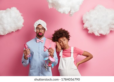 Tired Exhausted Pregnant Woman Leans At Shoulder Of Husband Who Stands With Baby Items, Become Parents Soon, Isolated On Pink Wall, Clouds Overhead. Future Father Holds Mobile, Feeding Bottle, Diaper