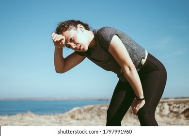 Tired Exhausted Young Athletic Woman Runner Wipes Sweat From Forehead, Taking A Rest, Doing Break, Breathing Hard After Running Hard At Mountain Landscape Nature Path Near Blue Sea In Hot Sunny Day